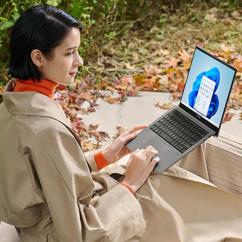 A woman sitting on the ground in a forest holding an ASUS Zenbook S 13 OLED laptop on her laps
