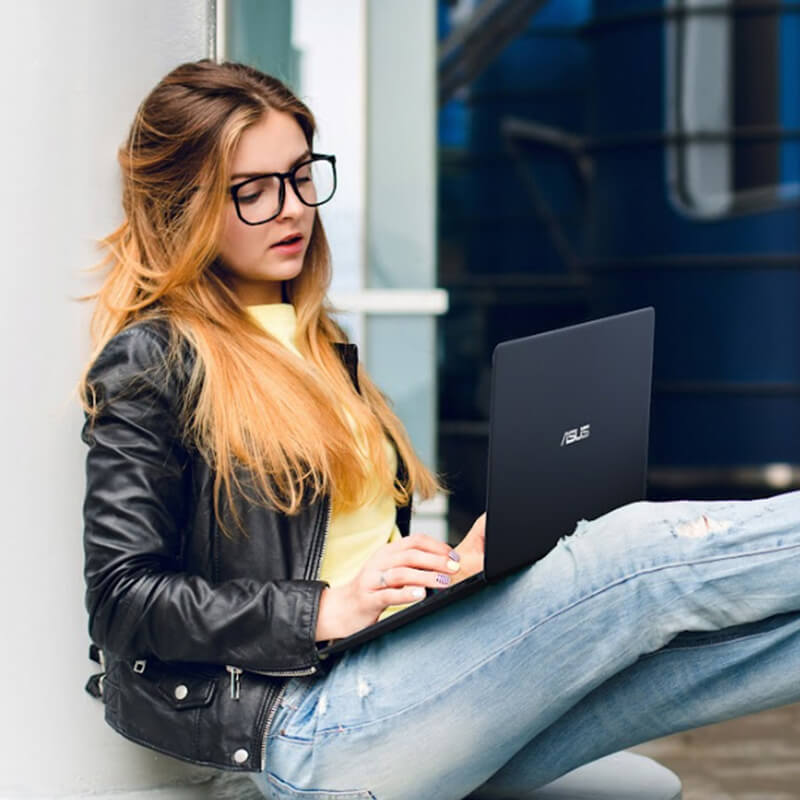 a young woman traveler uses ASUS Zenbook laptop while sitting at the airport with her legs on her suitcase