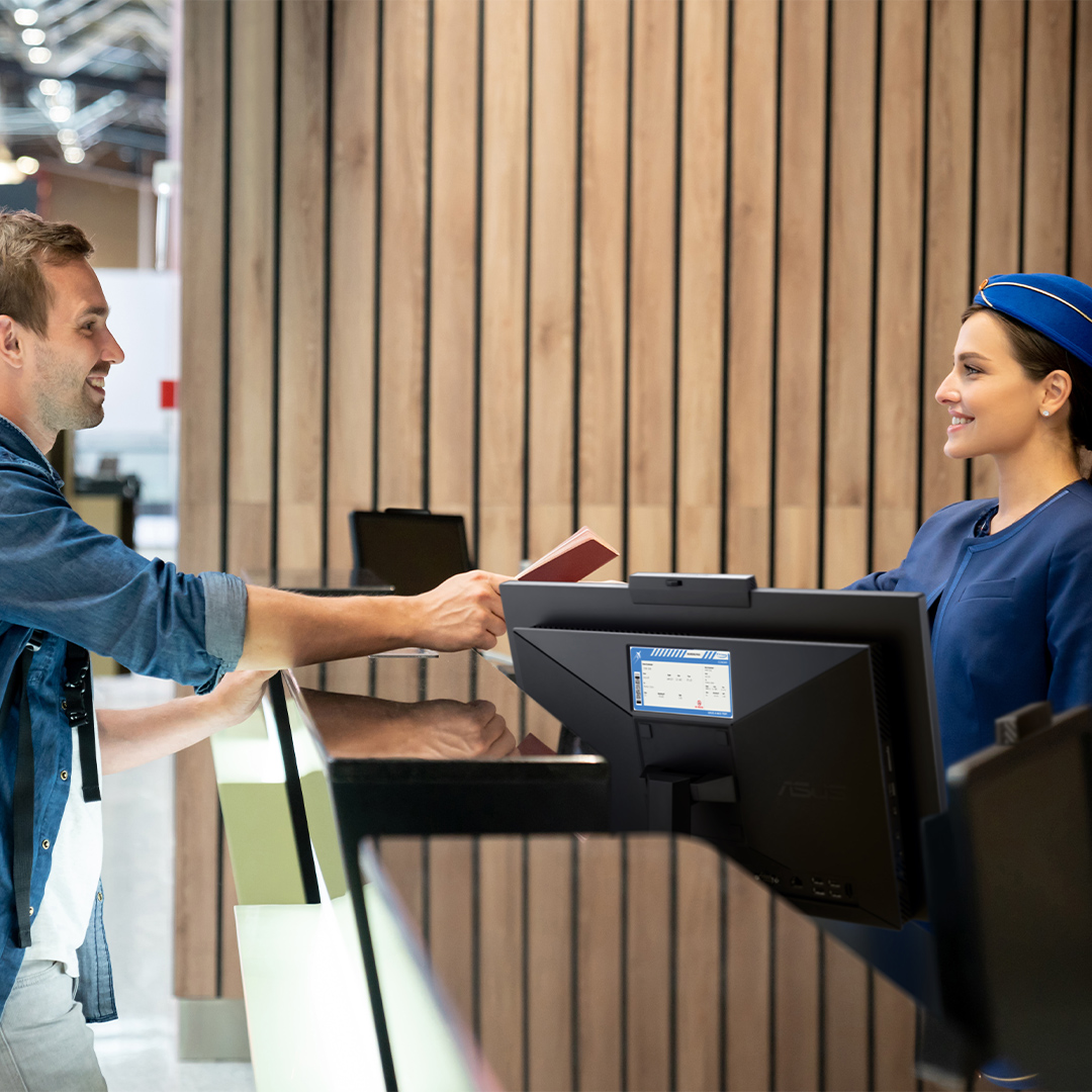 男子在機場櫃檯辦理登機手續，將護照交給身穿制服的女性地勤人員，照片中展示出ASUS ExpertCenter AiO 電腦背面的第二面顯示器