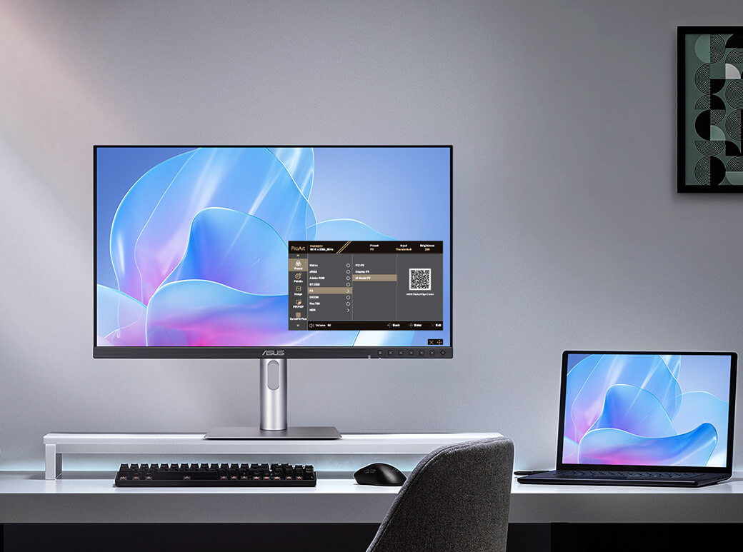 A modern desk setup with a ProArt monitor and a laptop displaying the same abstract blue and pink design. A keyboard and mouse are on the desk. The ambiance is sleek and minimalist.