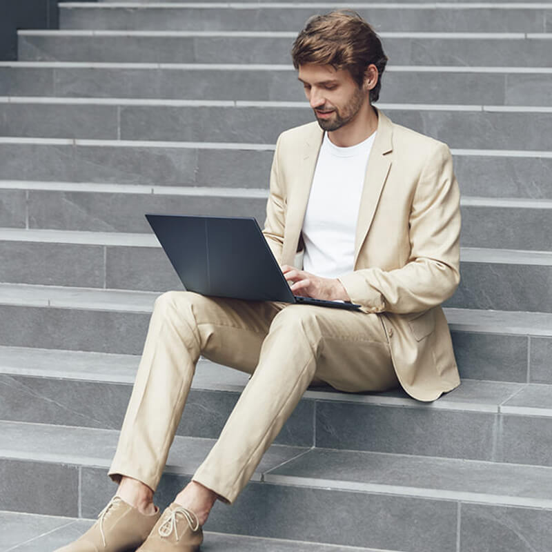 A man in a beige suit is using ASUS Zenbook 14 OLED on his lap while sitting on grey stone stairs.