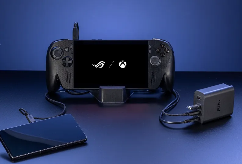 An ROG Xbox Ally X, connected to an ROG 100W Gaming Charger Dock and an ROG Phone, on a dark blue table.