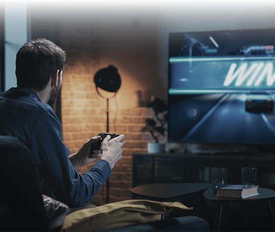 Un uomo tiene in mano un controller e gioca a un videogioco di corse su un grande schermo televisivo che mostra la parola “WIN” in un salotto moderno.