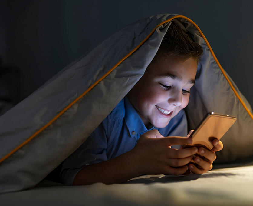 A school-aged boy is using a smartphone with a blanket covering his head.