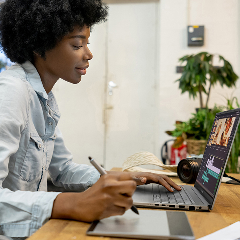 a young female content creator using pen tablet to edit videos on her ASUS Vivobook Pro 15 OLED creator laptop while sitting at a wooden table with a camera behind the laptop