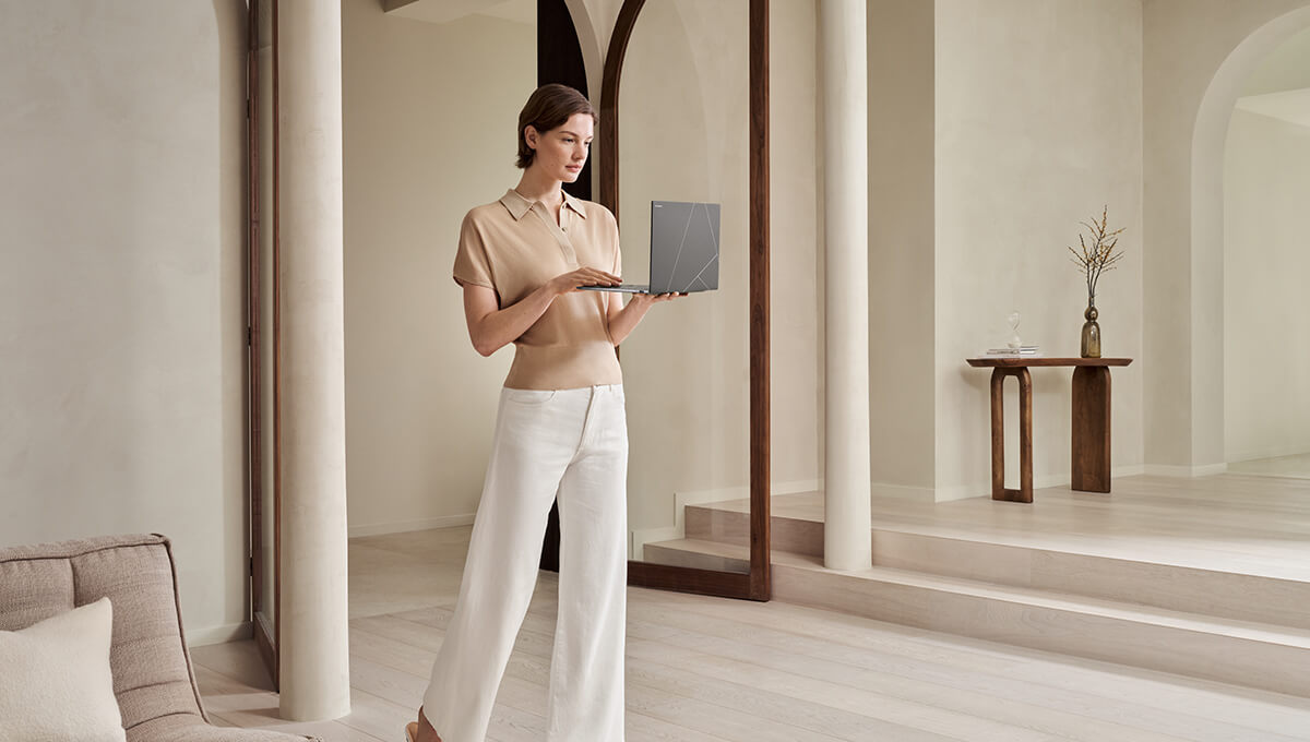 A young woman standing in a light-filled living room holding an open Zenbook S 14 laptop in her hands