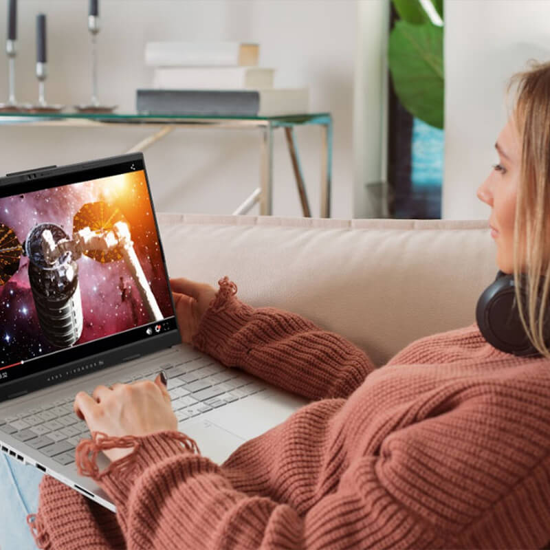 A young woman sitting on a pink sofa with an ASUS laptop on her lap, watching an online video