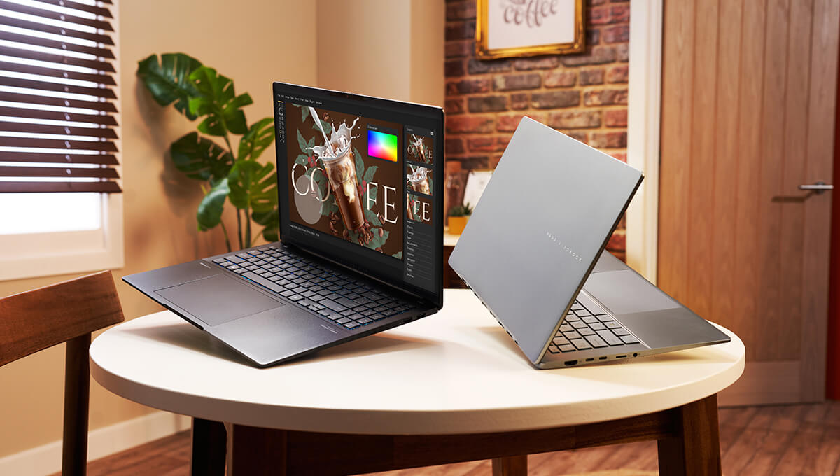 two open ASUS Vivobook S14 laptops in Neutral Black and Cool Silver colors lying on a table in a warm-colored, well-lit room