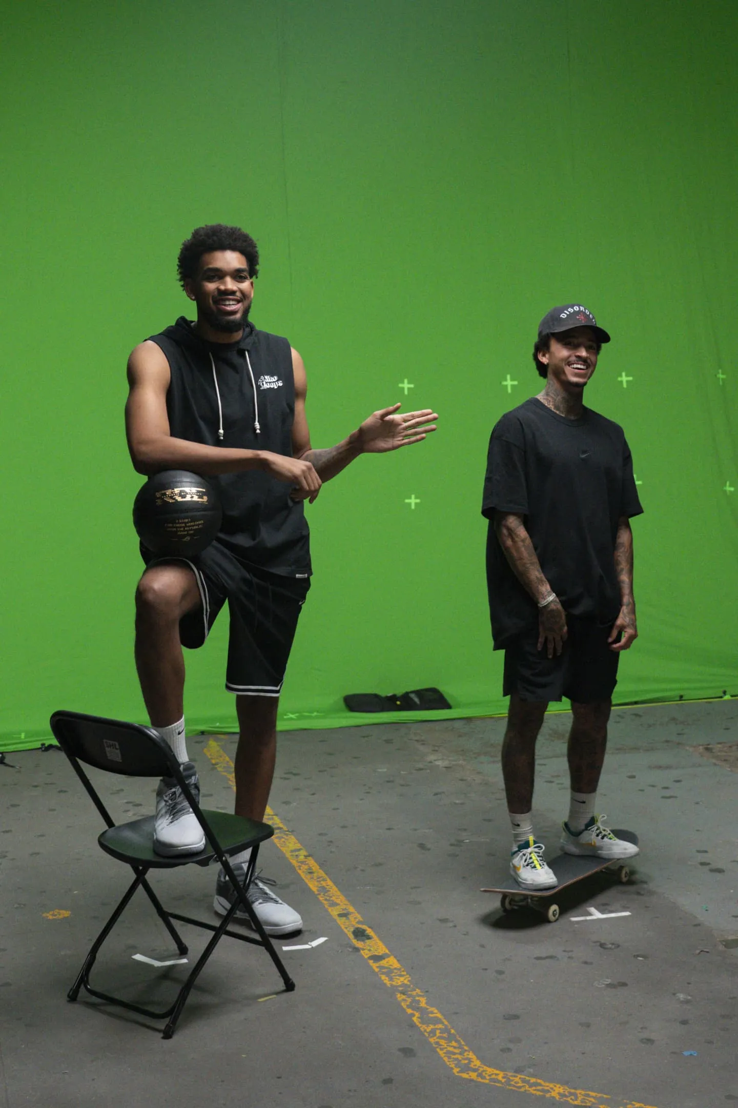 Karl-Anthony Towns and Nyjah Huston next to each other in front of a green screen, while Karl places his right leg on the chair and Nyjah is standing on a skateboard.