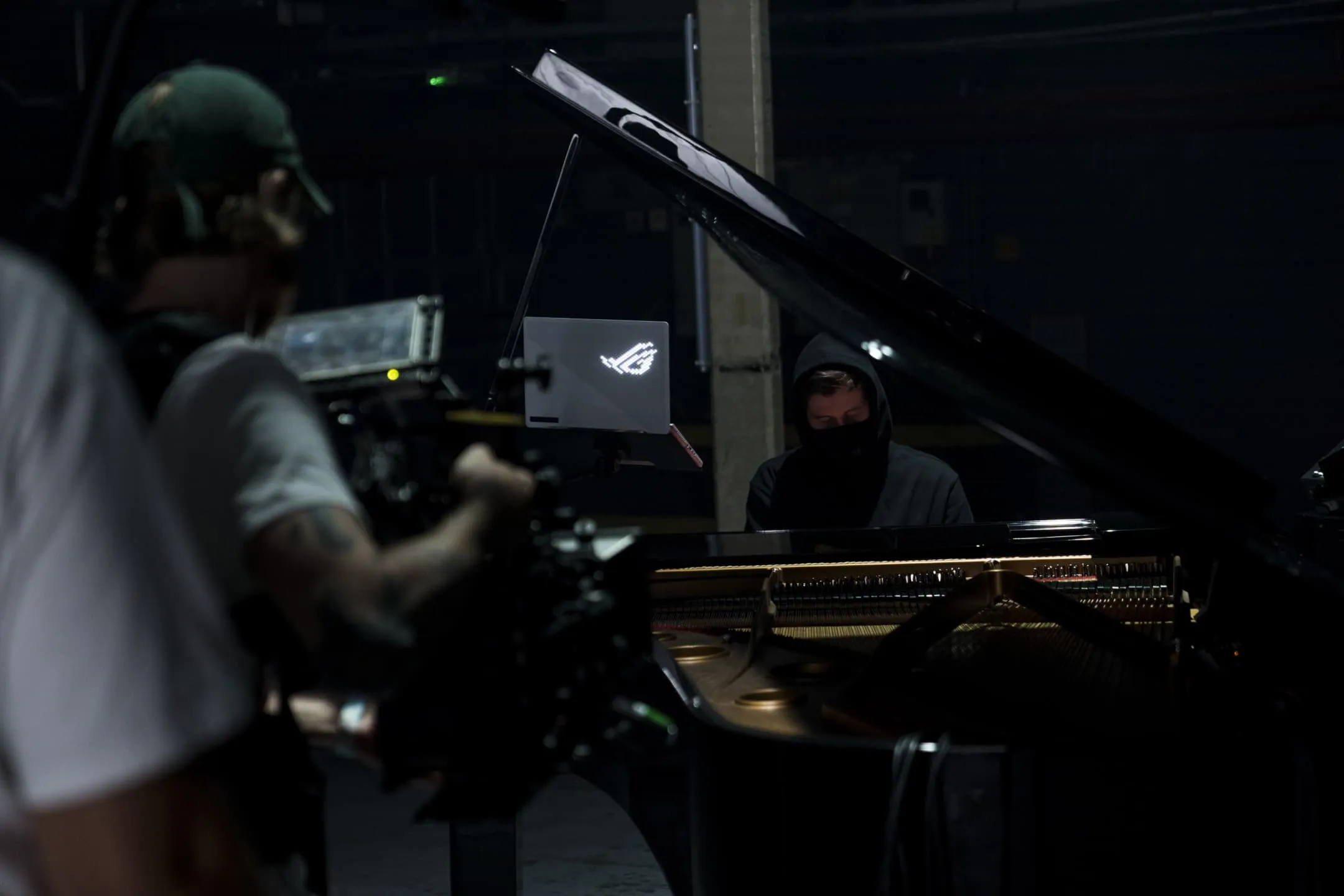 Alan Walker seated at a piano, with the Zephyrus G14 open nearby, and both being filmed by a camera crew in a warehouse.