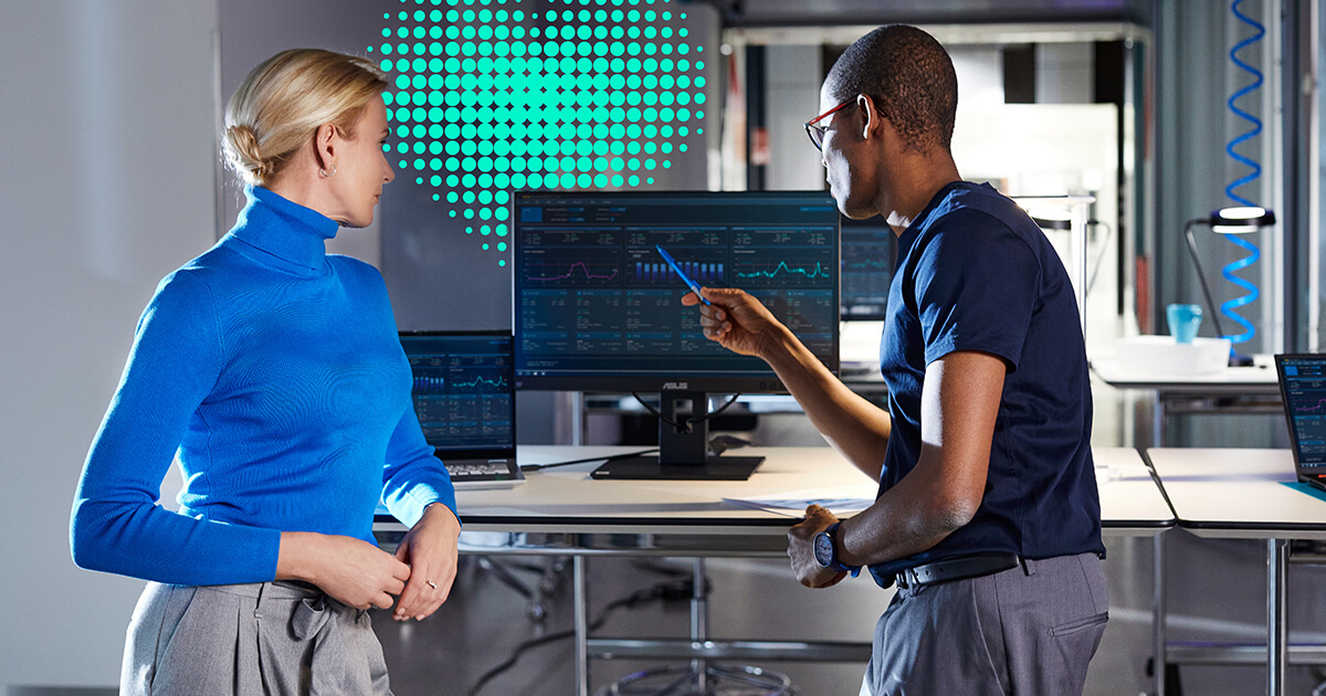 A woman on the left and a man on the right are discussing data displayed on the monitor between them. A green heart symbolizing care is hovering in the background.