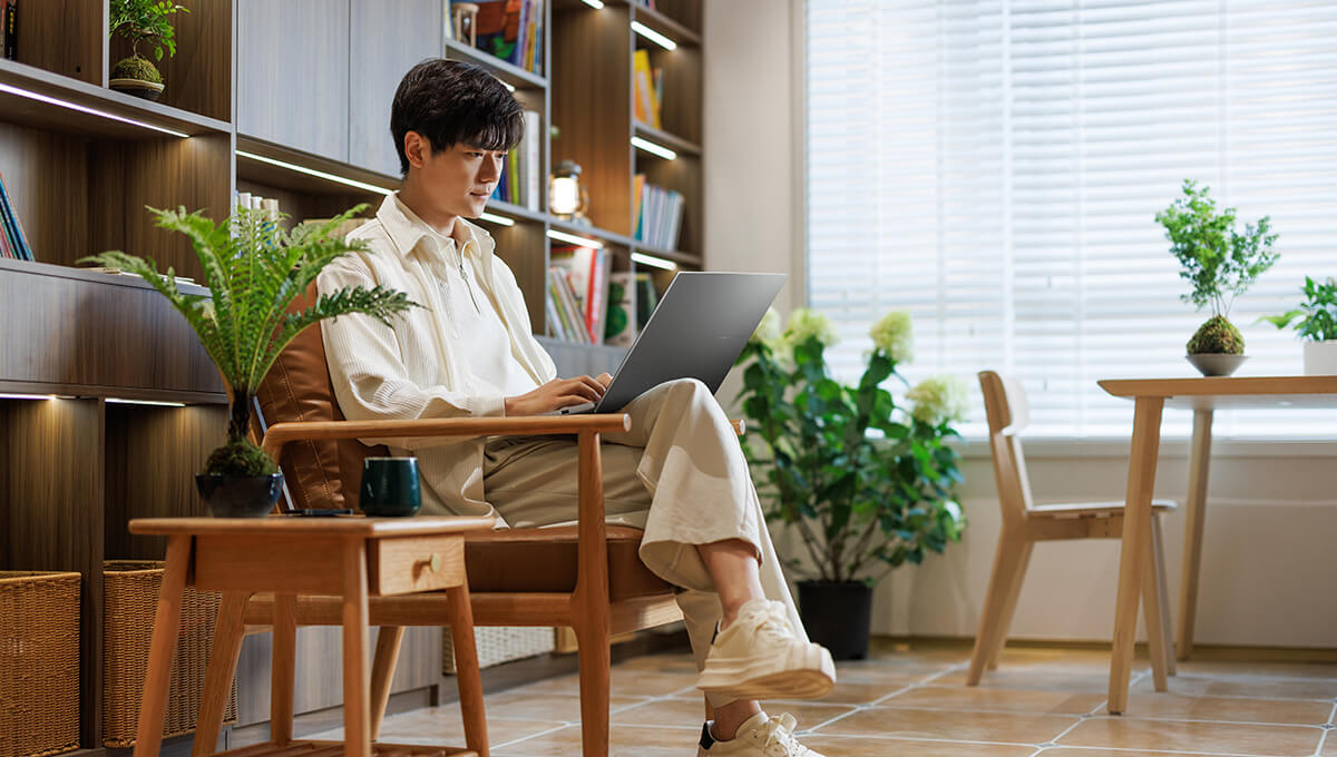 若い男性がASUS Vivobook S16を使用して椅子に座り、背景にいくつかの植物があります。