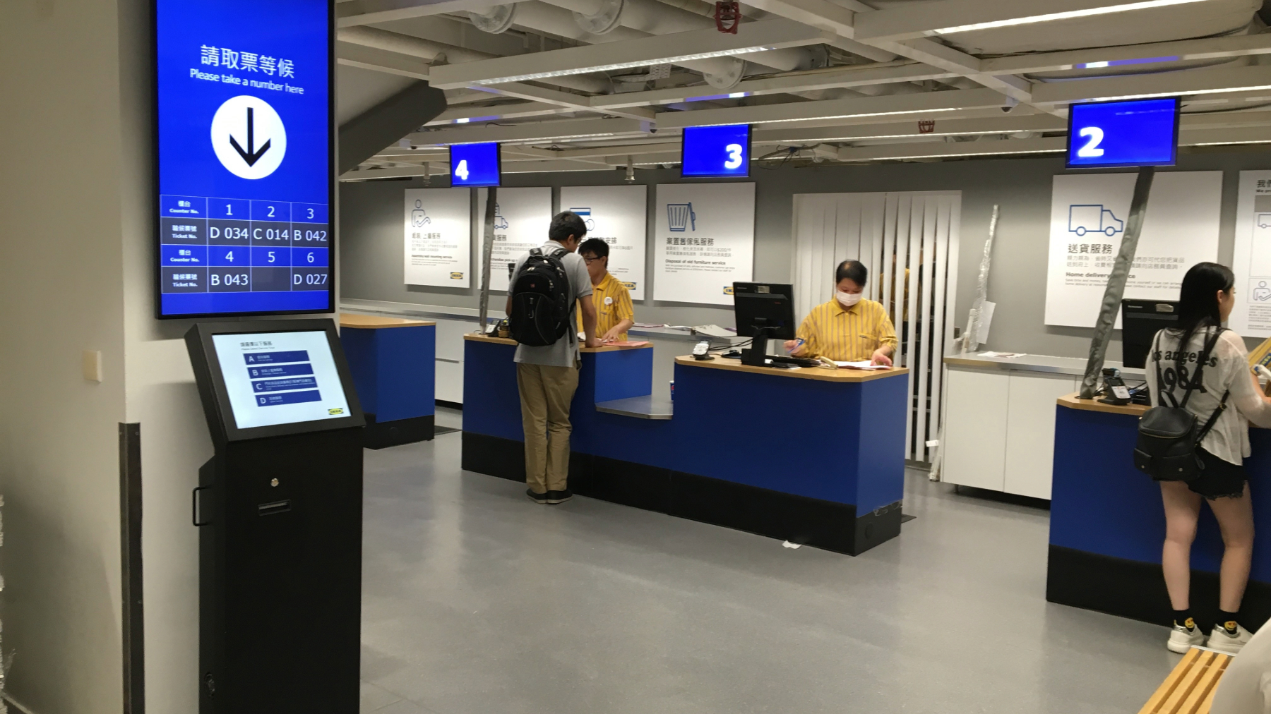 Interior of a service counter area with blue and white decor. A digital kiosk and screen display numbers. Staff in yellow uniforms assist customers.