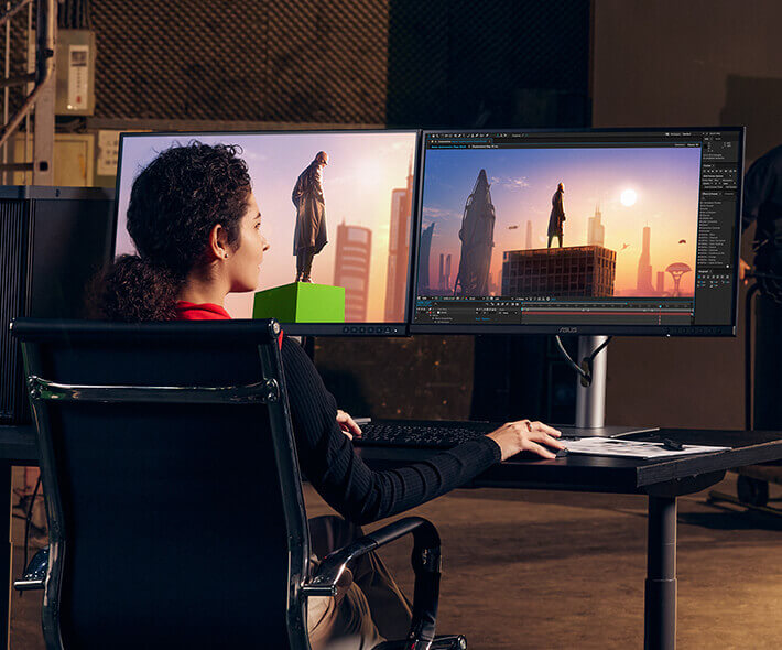A woman works with digital 3D modeling on dual monitors, featuring a futuristic cityscape and statue. The setting conveys creativity and focus.