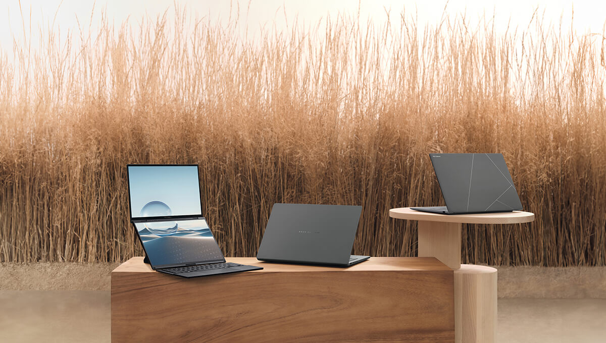 Three ASUS Zenbooks displayed on wooden stands in front of tall dry grass.