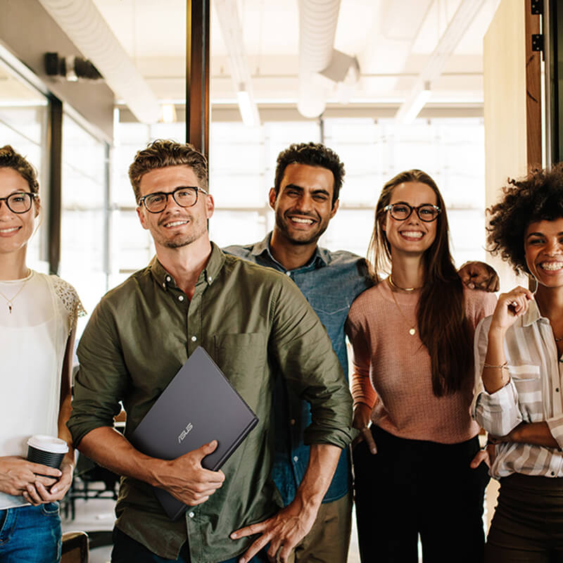 five smiling young professionals standing in a modern office environment with one of them holding a Zenbook Pro 16X OLED laptopn in one hand
