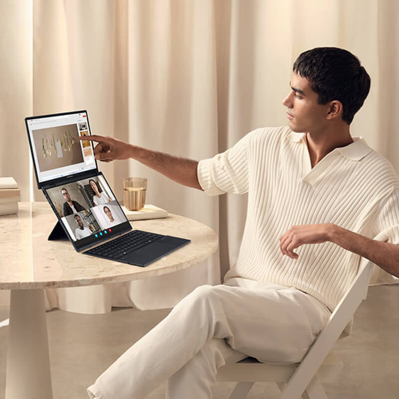 a young, brightly-dressed man touches the top screen of an ASUS Zenbook DUO laptop while displaying a Powerpoint presentation in a team meeting on the bottom screen