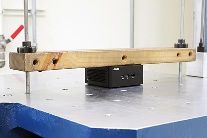 NUC Mini PC Vibration Test NUC best quality mini PC testing of vibration test, using a heavy wooden block applying pressure on the NUC mini PC as part of a stress or compression test on a metal surface.