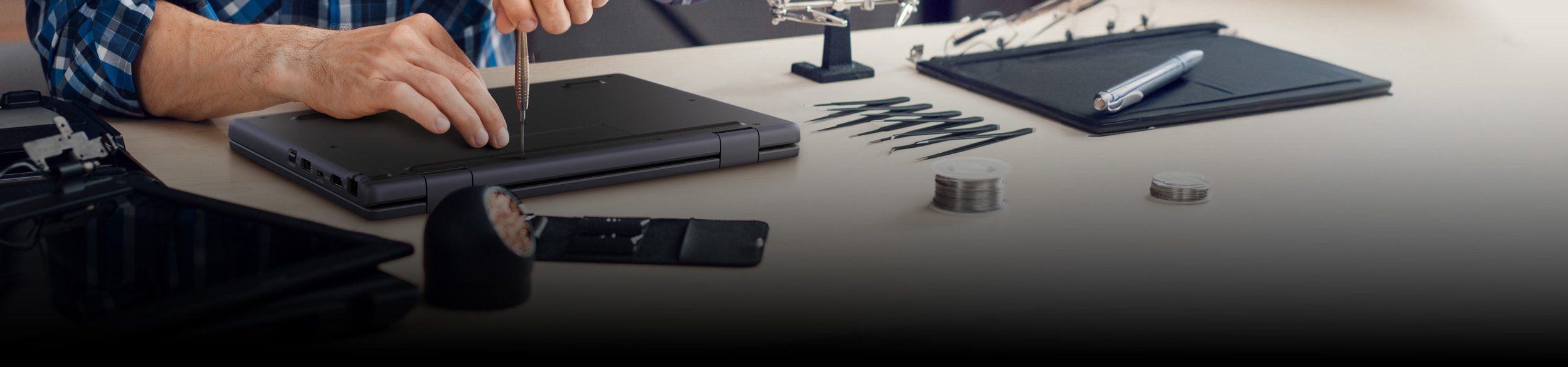 Une personne répare un ordinateur portable sur un bureau, entourée de tournevis de précision, de bobines de fil, d’un fer à souder et d’accessoires électroniques.