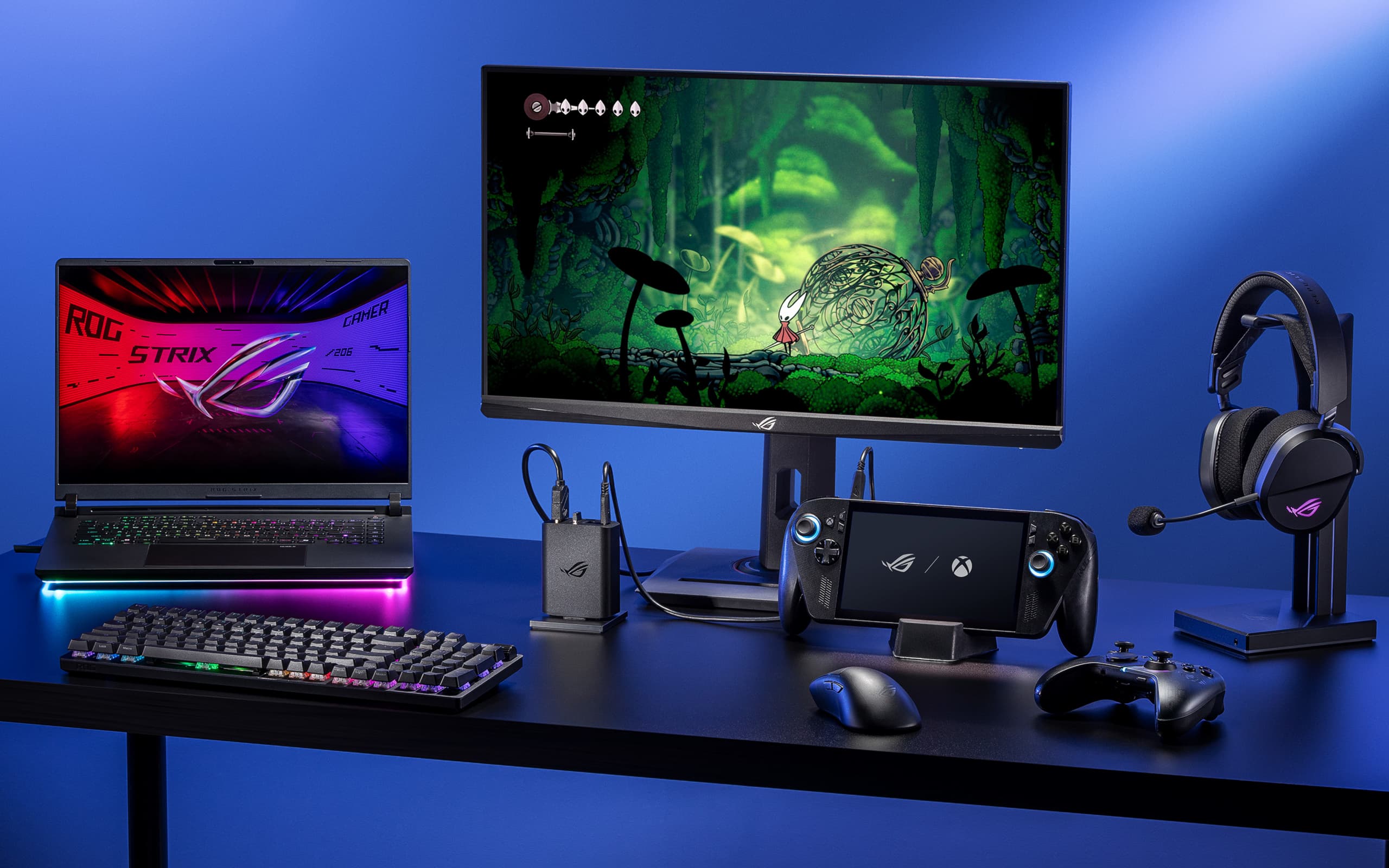 An ROG 100W Gaming Charger Dock plugged into a power port in the center of a desk, connected via HDMI to a monitor, and USB-C to an ROG Xbox Ally X. The desk also has an ROG Strix SCAR 16 laptop, wireless keyboard, wireless mouse, and an ROG headset on display.