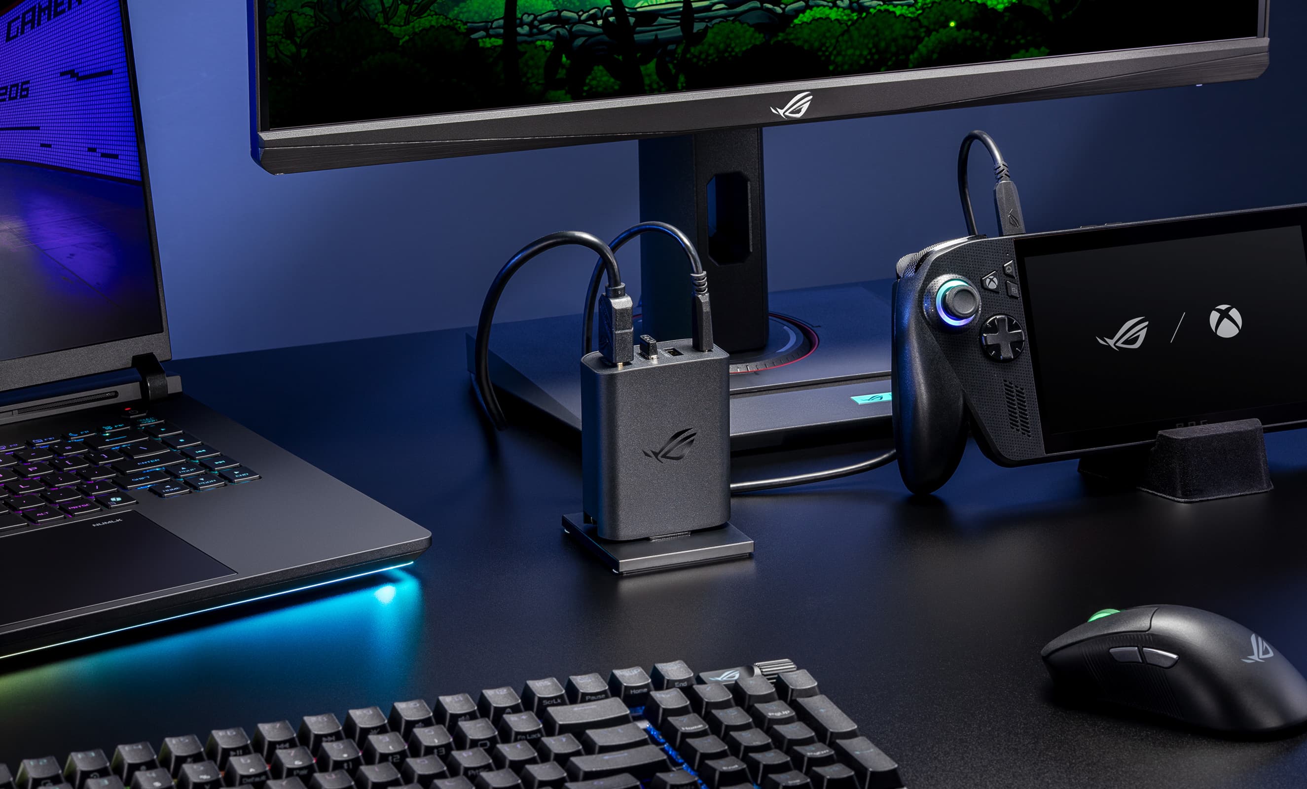 A close-up view of an ROG 100W Gaming Charger Dock plugged into AC power in the center of a desk, connected to a monitor and an ROG Xbox Ally X, with an ROG wireless mouse and keyboard, as well as an ROG Strix SCAR 16 laptop visible in the foreground.