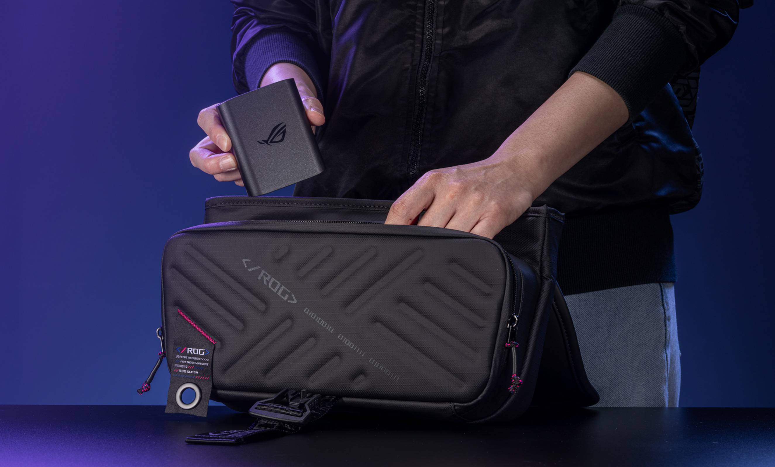 A person in a black jacket slipping the ROG 100W Gaming Charger Dock into an ROG Slash Sling Bag 4.0.