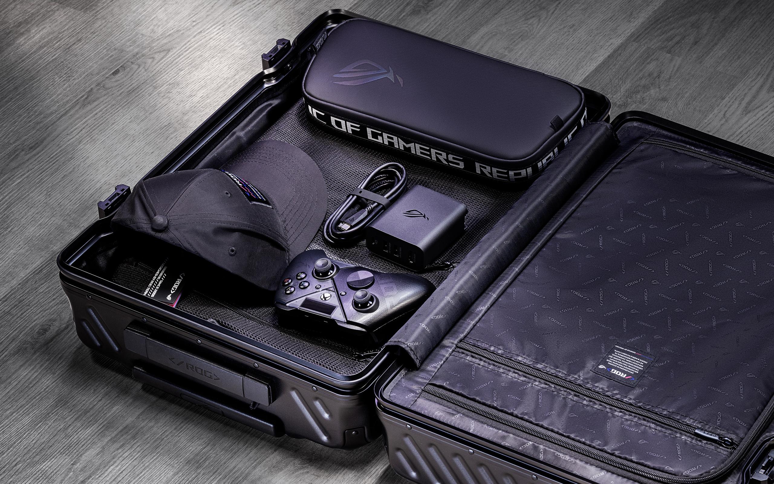 A black piece of luggage is open on the floor, with an ROG Ally X carrying case, an ROG cap, a Raikiri Pro PC controller, and an ROG 100W Gaming Charger Dock and its USB-C cable visible on one side of the open luggage.