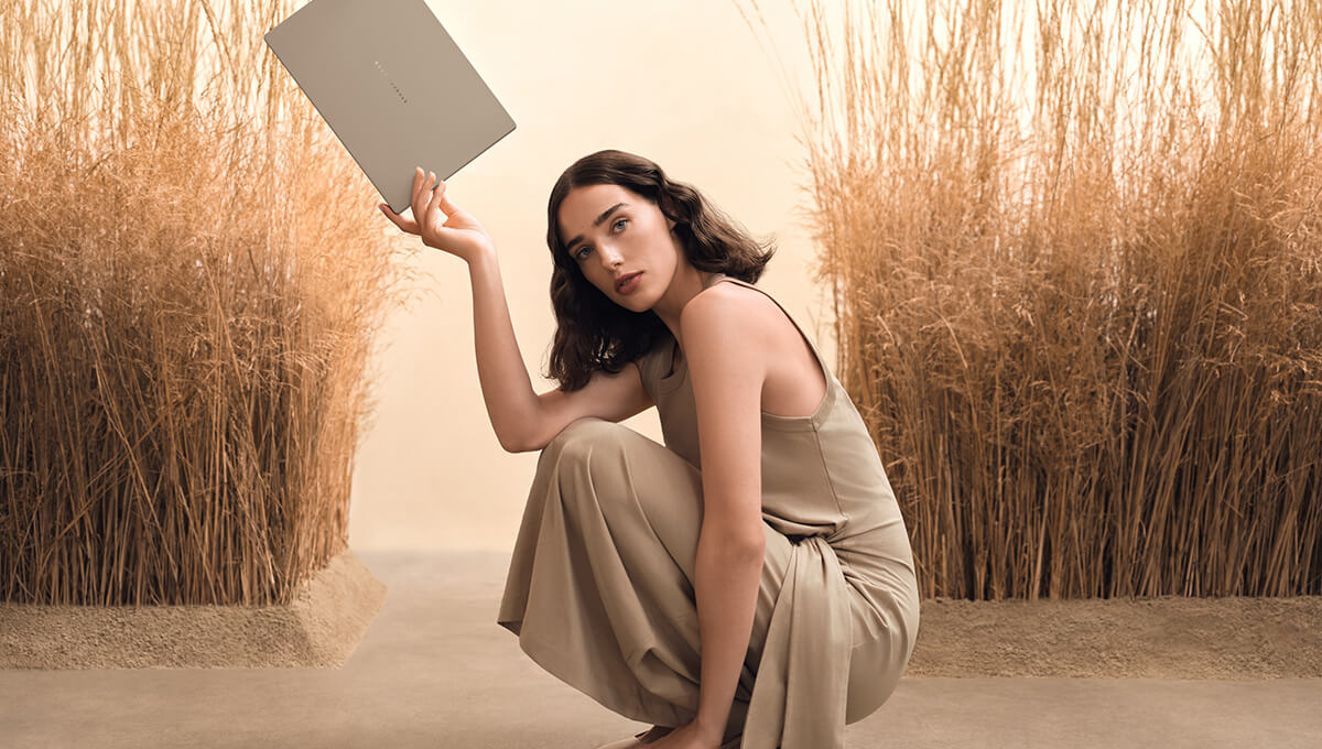 A woman in beige is crouched on the floor while she balances an ASUS ASUS Zenbook A14 between her fingertips.