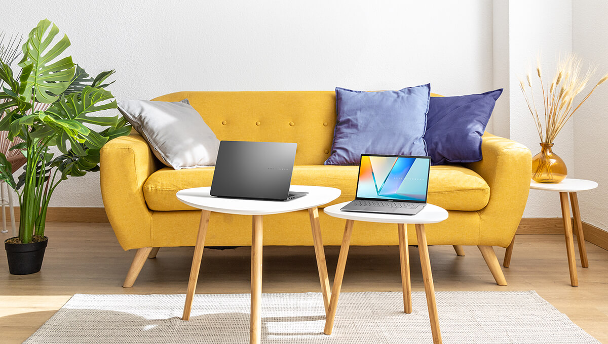 Two ASUS Vivobooks are placed on white triangular tables in the center of a bright, airy living room setting.