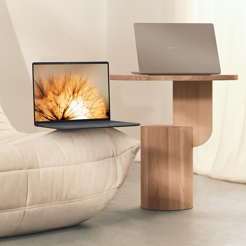 Two ASUS ASUS Zenbook 14 laptops sit open in a modern-looking room, one on the edge of a sofa and one on a wooden table beside the sofa.