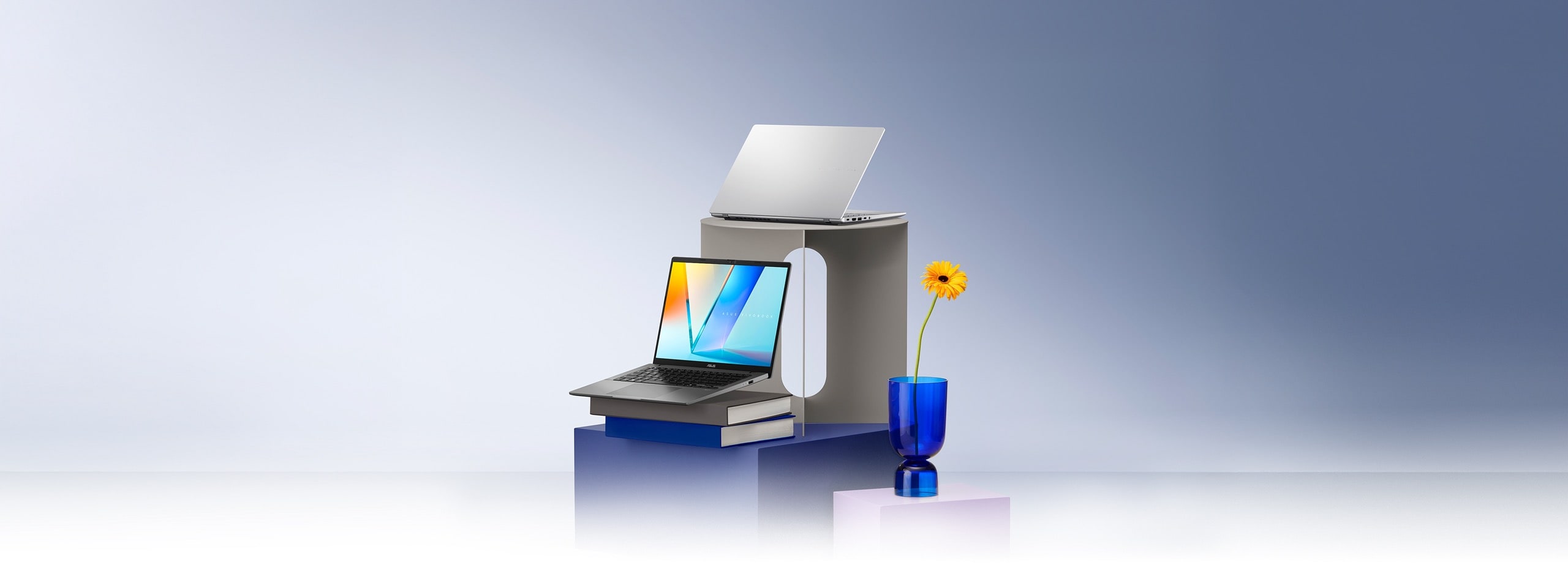 Two open laptops, one gray and one silver, are displayed on stacked gray, blue, and purple pedestals, with one resting on a pair of books. A single yellow flower in a blue glass vase stands nearby.