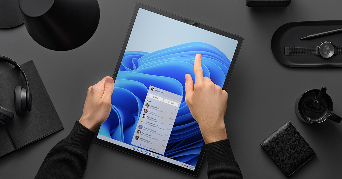 A person using a single hand to hold and a finger to control the touchscreen of the Zenbook 17 Fold OLED with Windows 11 fully extended in Tablet Mode on the desk with elegant black lamp, headphones, watch, clock, pen, cup of coffee and a wallet.