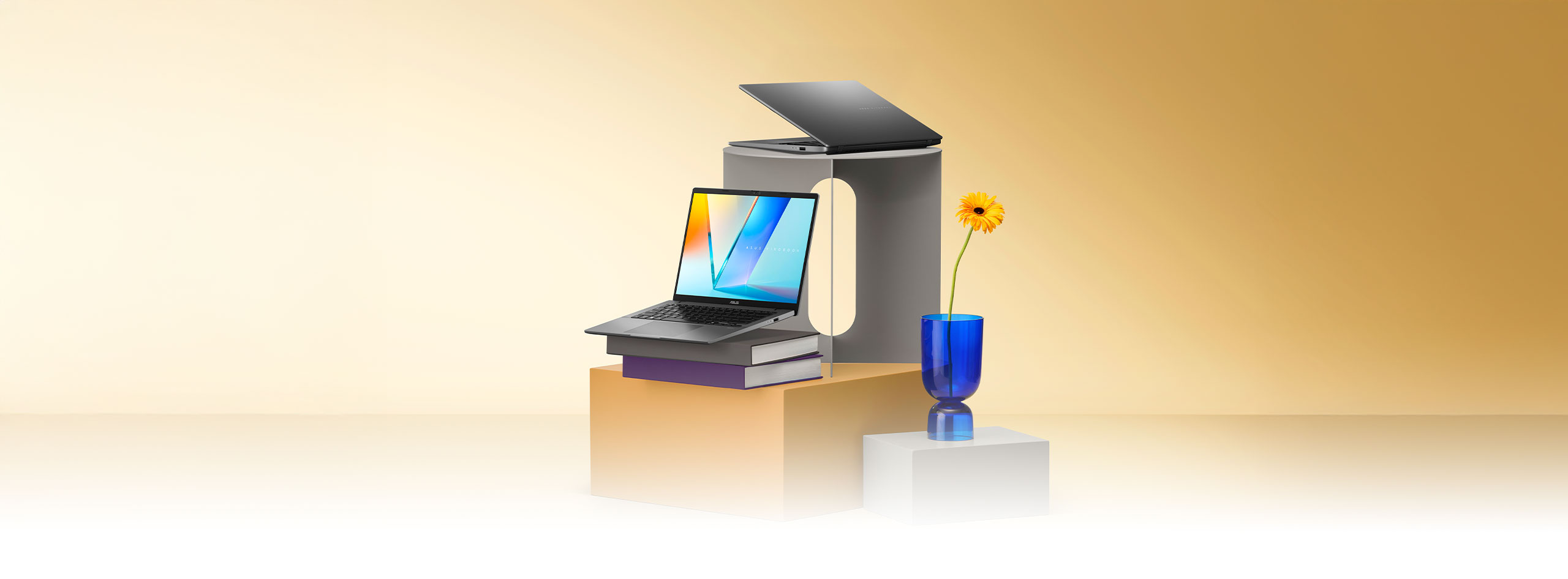 Two open laptops, one gray and one silver, are displayed on stacked gray and orange pedestals, with one resting on a pair of books. A single yellow flower in a blue glass vase stands nearby.