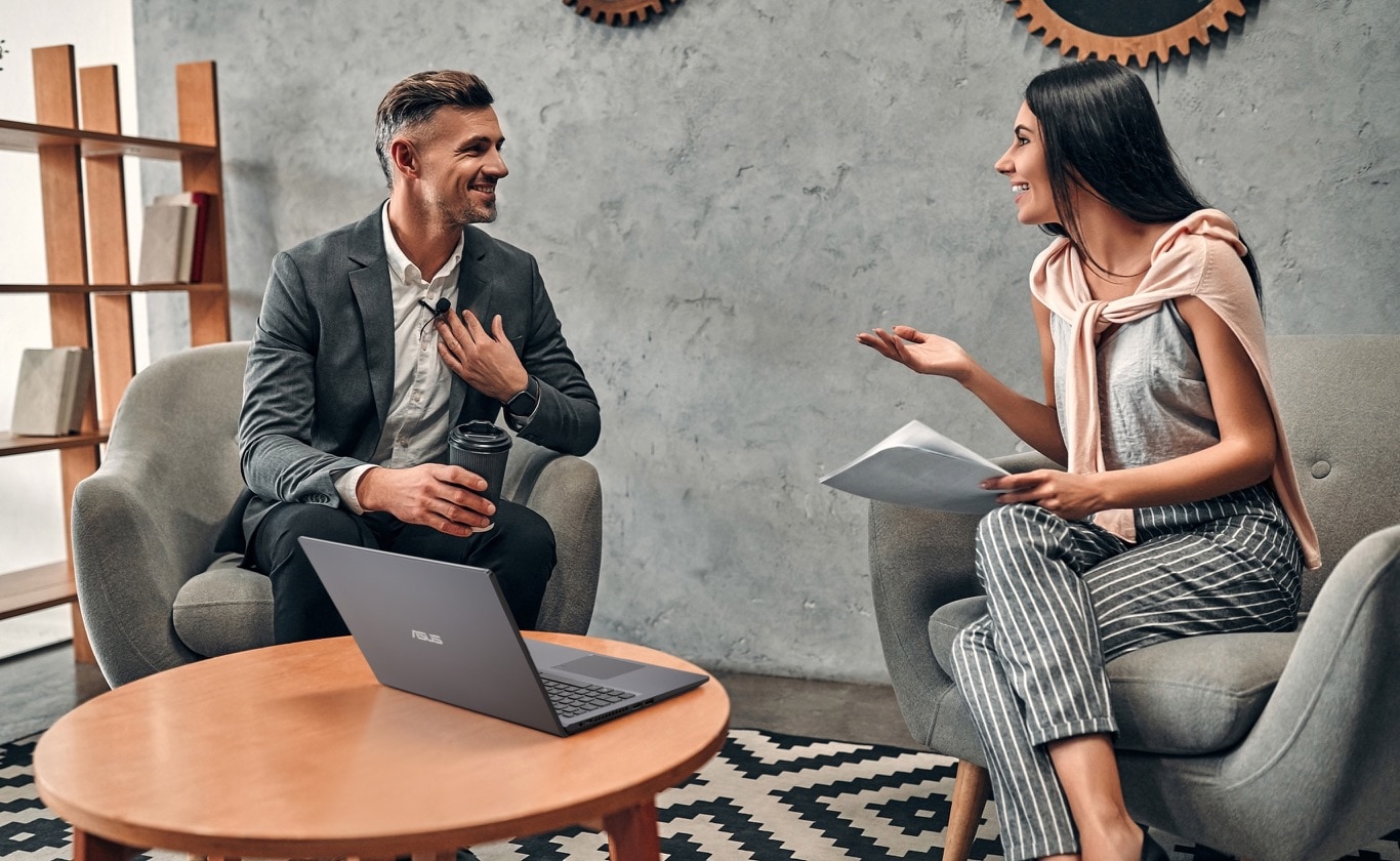 A man holding a cup of coffe is talking to a woman holding the documents. Both of them are sitting on the chairs and having good conversation. P1512 is put on the desk. 