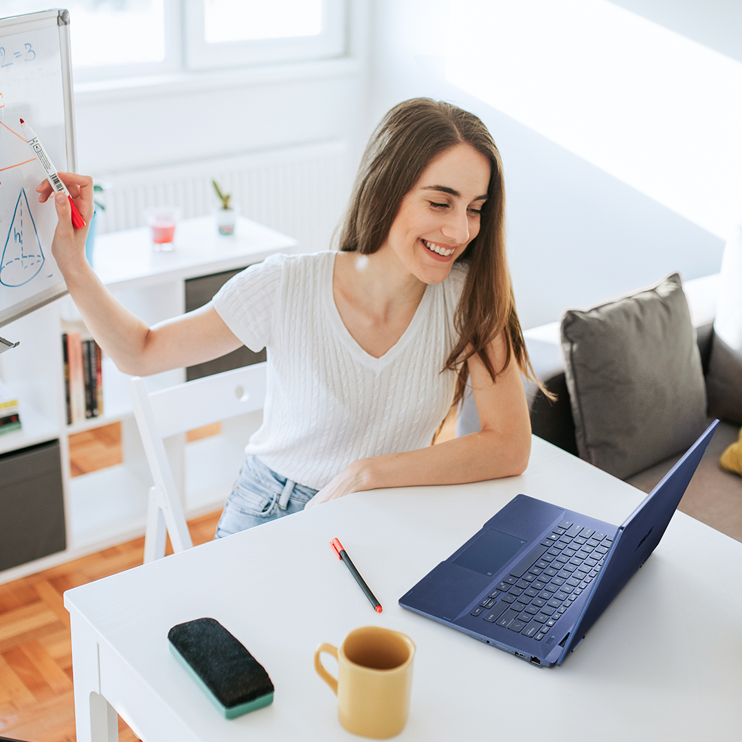 A teacher is looking at  ASUS ExpertBook B3 Flip to give remote lesson in her room, while one hand is writing on a whiteboard behind her.