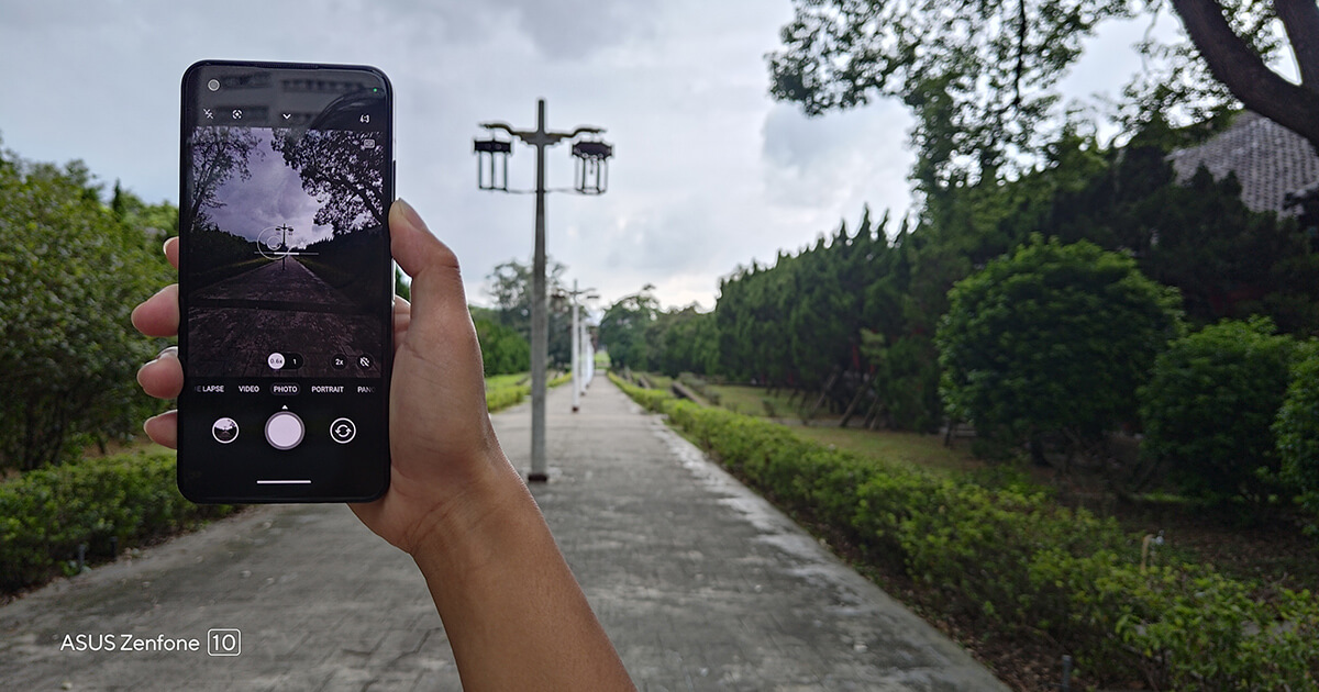 A zenfone 10 held with a right hand capturing a scenery of a long path with greenery on both sides and elegant streetlamps lined down the middle.