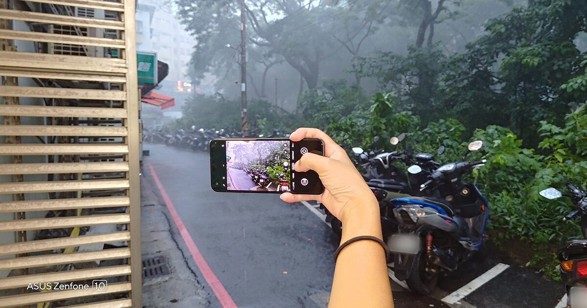 a person holding a Zenfone 10 in one hand horizontally while recording the rainy weather landscape, with some motorcycles in the background.