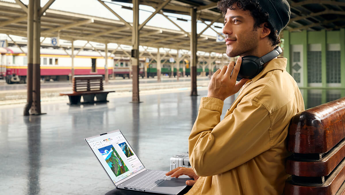 A man in a yellow shirt smiles as he looks to the left with one hand on his headphones around his neck and the other on the ASUS Vivobook on his lap.