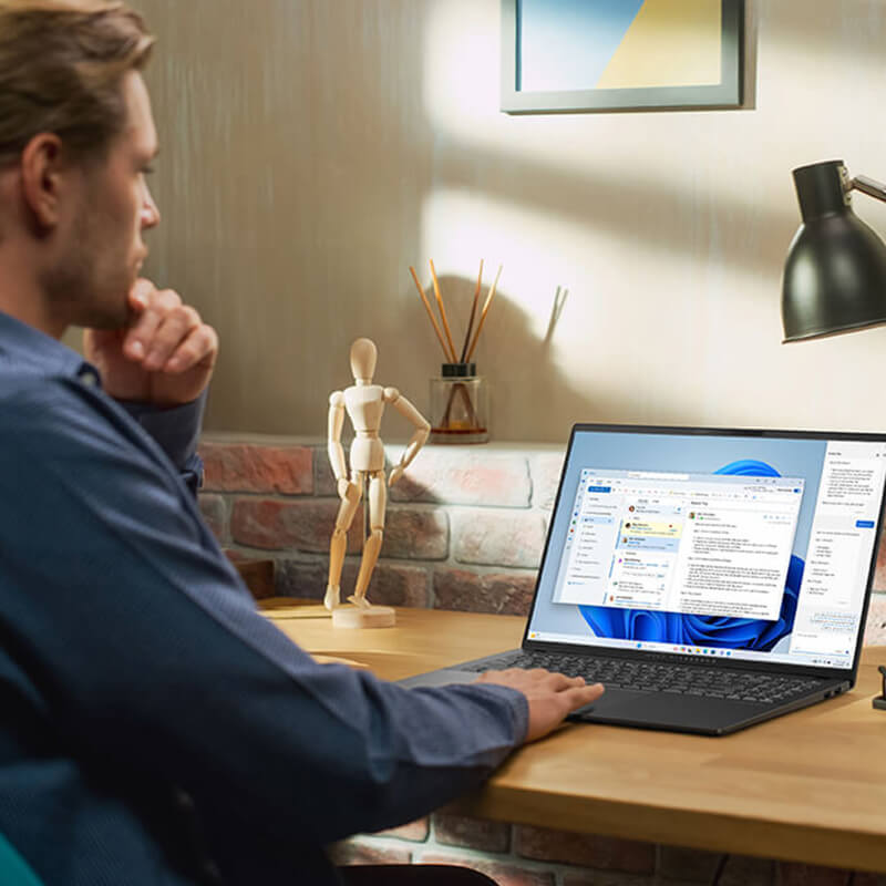 A man in a blue shirt is using Copilot features on an ASUS laptop placed on a wooden desk.