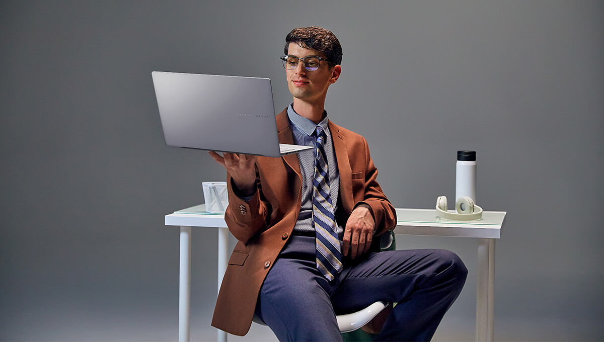 man in a suit holding an ASUS Vivobook S series laptop while sitting against a white table.