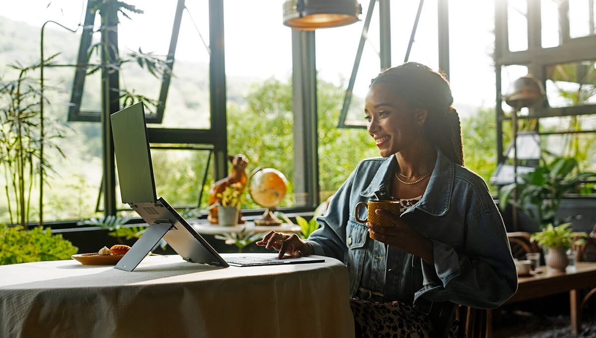 Una mujer joven con una sonrisa en su rostro mientras sostiene una taza, usa ASUS Zenbook DUO en modo de doble pantalla, sentada en una habitación llena de plantas