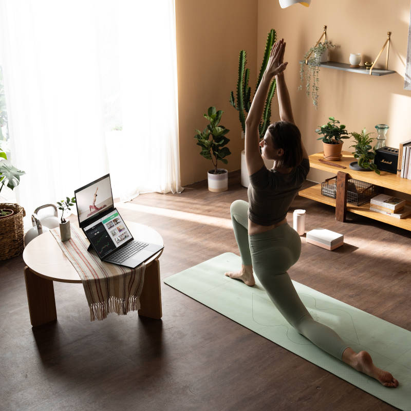 A la derecha, una mujer hace yoga sobre un tapete verde claro. Ella está mirando una laptop de doble pantalla apoyada en la mesa redonda a su izquierda.
                  Hay dos pantallas en la laptop. La de arriba muestra el tutorial de yoga de la mujer, mientras que la de abajo está en pantalla dividida y muestra información diferente.