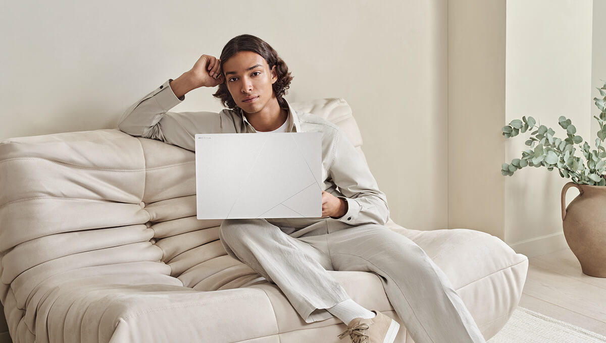 a young man sitting on a white sofa with a Zenbook S 16 laptop in Scandinavian White color on his lap
