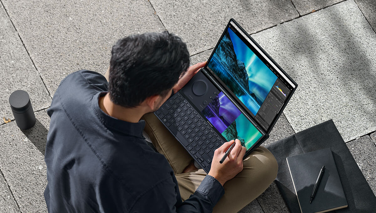 a top view of a young man using a stylus on the ASUS Screenpad plus of the Zenbook Pro Duo 14 OLED dual screen laptop while sitting on stairs outdoors