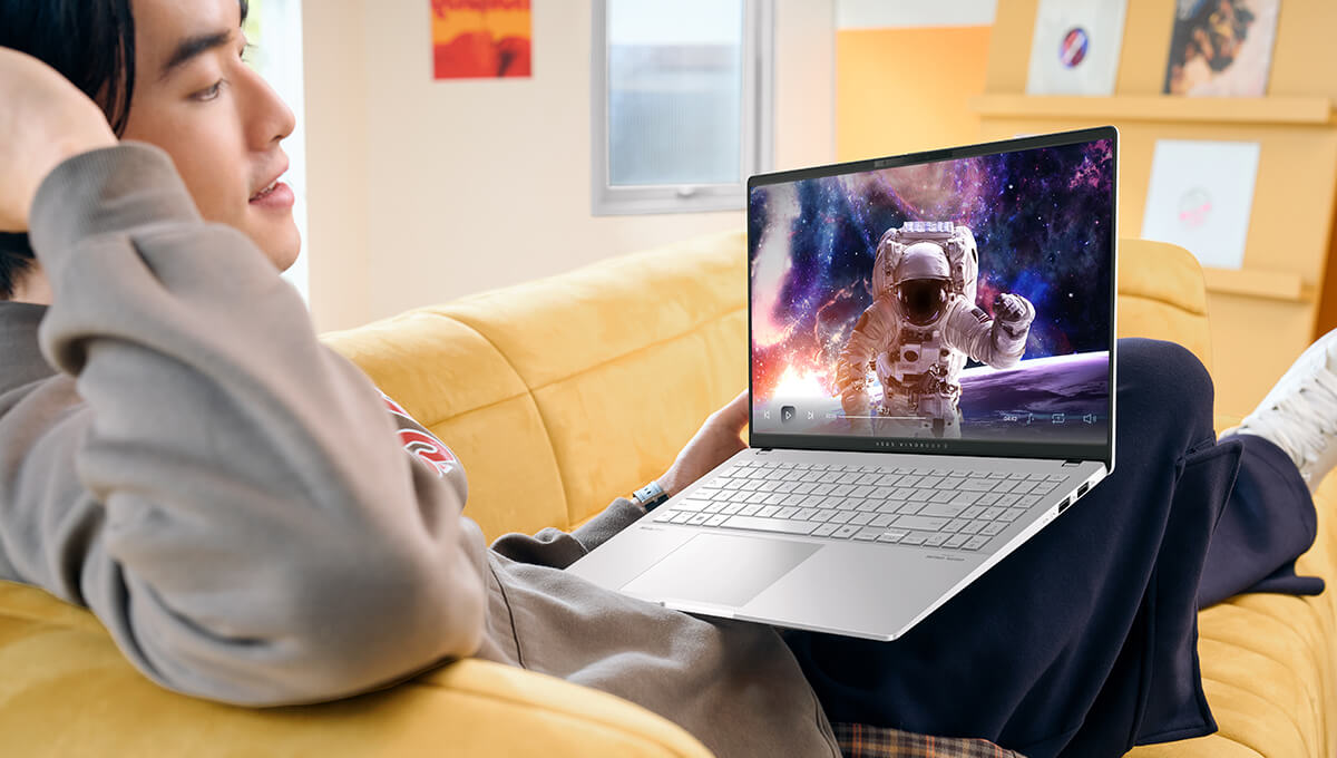 a man sitting on a sofa with his ASUS Vivobook S 15 laptop on his laps, watching a movie