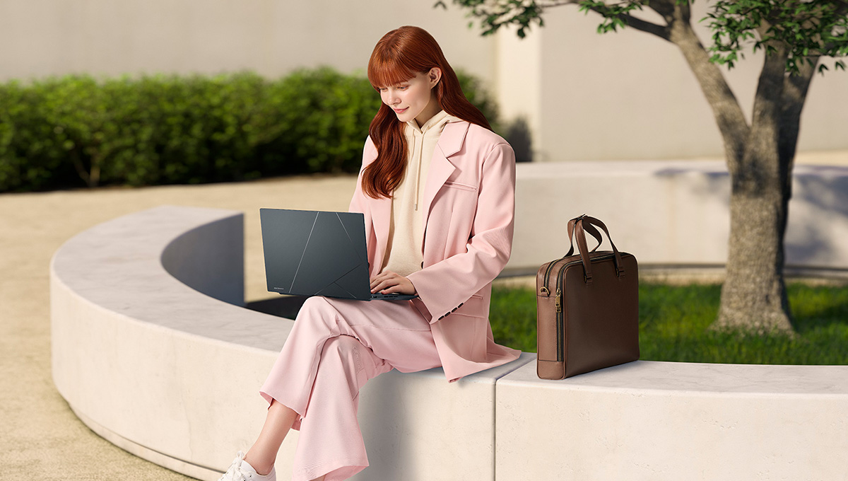 一名女士坐在低牆上，腿上放著一台 ASUS Zenbook 14X OLED 筆記型電腦，旁邊放著一個棕色公事包，背景是一棵樹
