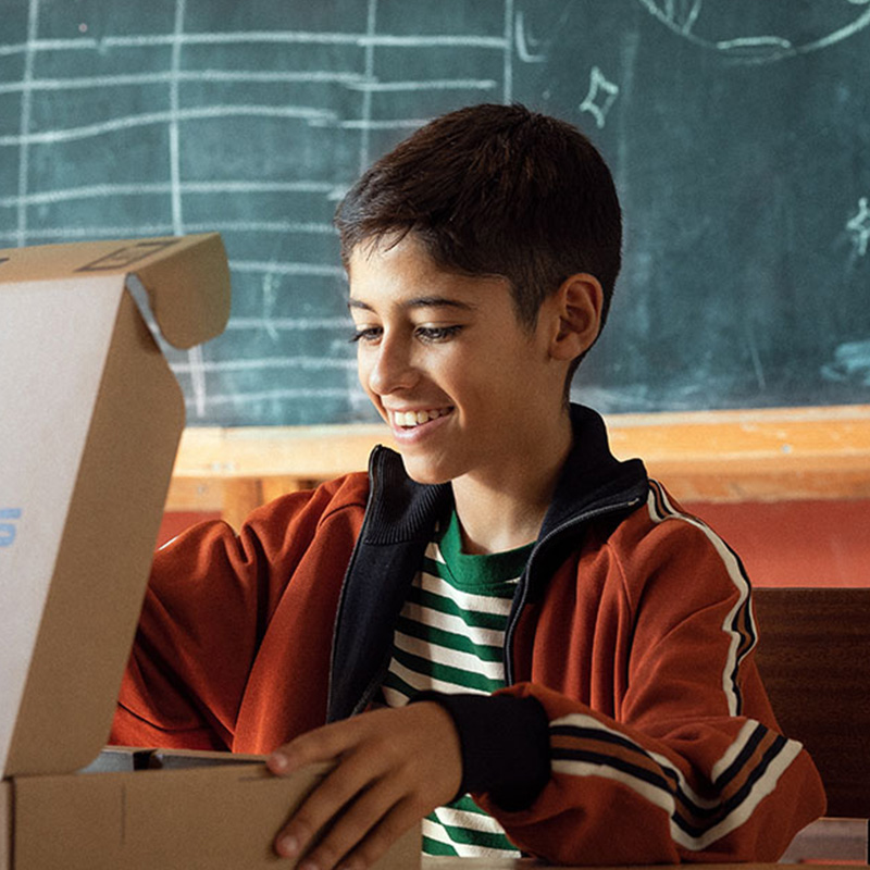 Un étudiant souriant ouvre une boîte d'ordinateur portable ASUS alors qu'il est assis devant un tableau noir dans une salle de classe.