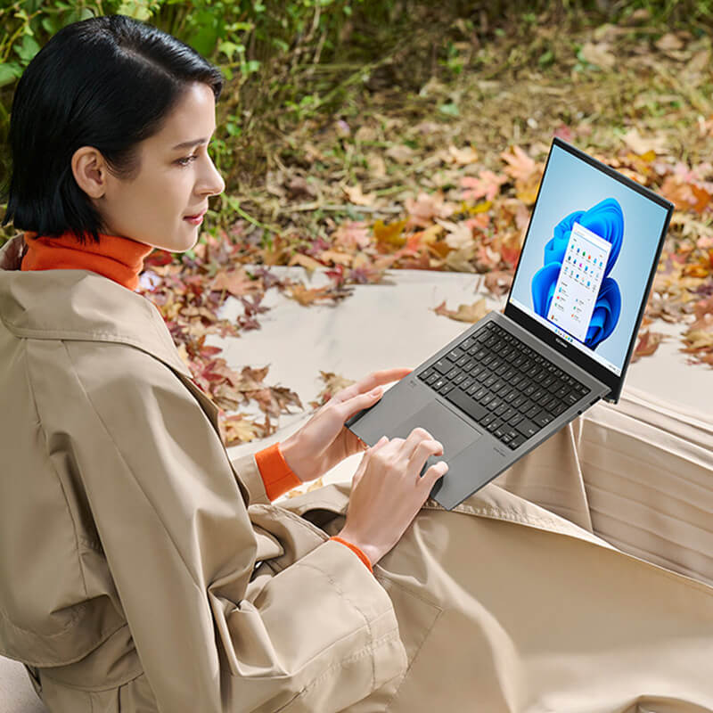 A woman sitting on the ground in a forest holding an ASUS Zenbook S 13 OLED laptop on her laps
