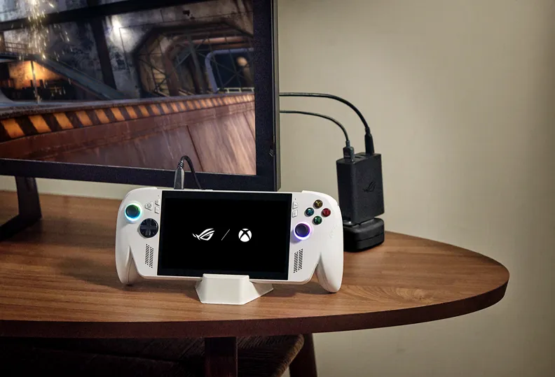 An ROG Xbox Ally X, connected to an ROG 100W Gaming Charger Dock and a TV on a wooden table.