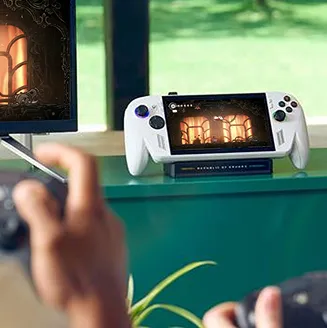 A person gaming with a controller in hand on a TV, with the ROG Xbox Ally sitting near the TV on a green counter.